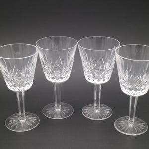 May include: Four clear crystal wine glasses with detailed cut glass patterns. Each glass features a stemmed base and a flared bowl. The glasses are displayed on a dark surface, highlighting their elegant design.