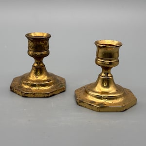Puede incluir: Dos candeleros antiguos de latón. Cada soporte tiene una base hexagonal y un tallo detallado que conduce a una parte superior en forma de copa. El latón tiene un aspecto envejecido y deslustrado, con desgaste y pátina visibles.