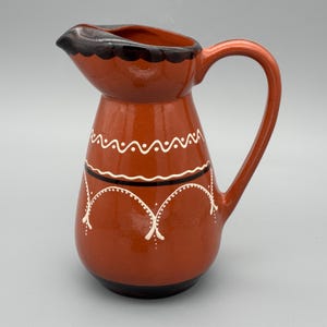 May include: A terracotta-coloured jug with a curved handle and spout. The rim is dark brown, and the body is decorated with white and black painted designs. The jug has a rounded base and a smooth, glossy finish.