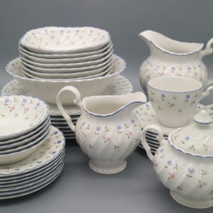 Könnte beinhalten: Ein Set aus weißem Porzellangeschirr mit floralem Muster. Das Set umfasst Teller, Schalen, einen Sahnekännchen, eine Zuckerdose und eine Teetasse. Jedes Stück hat einen blauen Rand und ist mit kleinen rosa und blauen Blumen verziert.