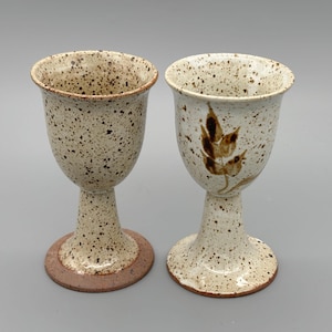 Puede incluir: Dos copas de cerámica moteada. Una copa tiene una base marrón y la otra una base blanca. Una copa tiene un diseño de hojas marrones. Las copas tienen un acabado moteado blanquecino.