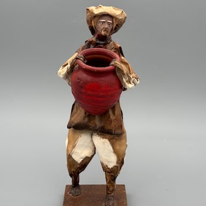 May include: A paper mache figurine of a man holding a large red pot. The figure is dressed in a brown outfit with white accents and a hat. The figurine stands on a small, rectangular base.