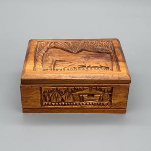 May include: A rectangular wooden box with intricate carvings. The lid features a landscape scene with trees and mountains. The front of the box has a carved design of a building and trees. The box is made of a light brown wood.