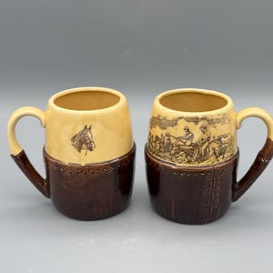 Puede incluir: Dos tazas de cerámica con la parte superior beige y la inferior marrón oscuro. Una taza presenta una cabeza de caballo, la otra una escena de carruaje. Ambas tienen asas marrones y un diseño cosido en la parte inferior.