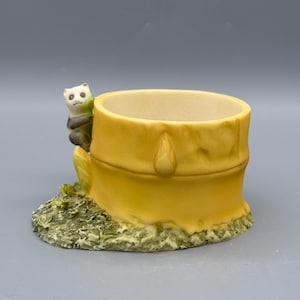 Può includere: Una statuetta in ceramica di un panda che si aggrappa a un contenitore giallo a forma di bambù. Il contenitore ha un interno chiaro e un esterno strutturato con un design a goccia. La base è verde con piccoli dettagli.