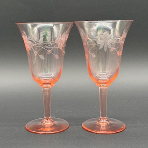 Peut inclure: Une paire de verres à pied en verre rose avec un motif floral gravé délicat autour du bol supérieur. Chaque verre possède une tige fine et une base circulaire, présentant une teinte rose translucide avec des gravures détaillées de feuilles.