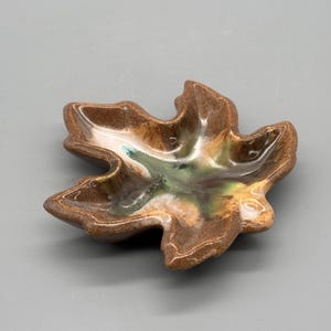 May include: A ceramic leaf-shaped dish with a glossy finish. The dish features a blend of brown, green, and cream colours. The edges are a darker brown, and the interior has a marbled effect. This decorative dish is ideal for holding small items.