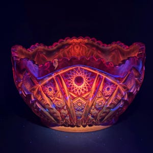 May include: A deep red glass bowl featuring a complex pressed pattern of geometric stars and diamond shapes. The bowl has a scalloped, serrated rim and a translucent quality that glows with vibrant red light against a dark background.