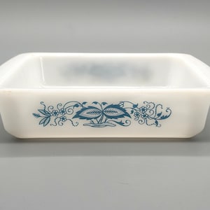 May include: A rectangular, off-white Pyrex baking dish with a blue floral design. The design features stylized leaves and flowers. The dish is empty and sits on a gray surface. A classic kitchenware item.