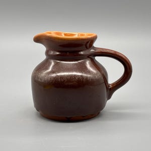 Peut inclure: Un petit pichet en céramique marron avec une anse et un bec verseur. L'intérieur du bec verseur est d'une nuance de marron plus claire. Le pichet a un corps arrondi et une finition lisse et brillante. La poignée est incurvée et fixée sur le côté du pichet.