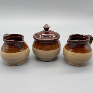 Könnte beinhalten: Ein dreiteiliges Keramikset mit einer Zuckerdose mit Deckel, einem Sahnekännchen und einem kleinen Krug. Die Teile sind braun und beige mit glänzendem Finish. Die Zuckerdose hat einen kleinen Knopf auf dem Deckel.