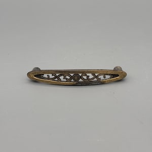Peut inclure: Une poignée de tiroir de style antique, de forme ovale, avec un motif de filigrane floral décoratif. La quincaillerie est en métal de couleur bronze et présente un aspect légèrement vieilli. La poignée mesure environ 13 cm de long.
