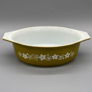 May include: A round, olive green casserole dish with integrated side handles and a white interior. The exterior features a repeating white floral pattern consisting of stylised blossoms and small dots encircling the entire circumference.