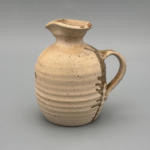 Puede incluir: Una jarra de cerámica beige con cuerpo redondeado, asa y pico. La jarra tiene una textura moteada y estrías horizontales. Diseños marrón oscuro en forma de rama decoran el lateral. La jarra es probablemente hecha a mano.
