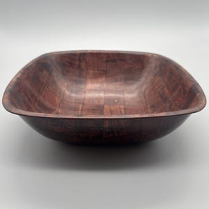 Puede incluir: Un cuenco cuadrado de madera con esquinas redondeadas. El cuenco está hecho de madera marrón oscuro con un patrón de vetas visible. El interior y el exterior del cuenco son del mismo color y textura.