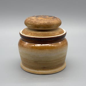Peut inclure: Un pot en céramique avec un couvercle. Le pot est de couleur beige clair avec des rayures horizontales marron. Le couvercle est marron foncé avec un motif circulaire sur le dessus. Le pot est probablement utilisé pour le stockage.