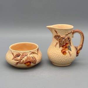 Op de afbeelding: Een crèmekleurige keramische melk- en suikerpotset. De set heeft een getextureerd oppervlak en is versierd met bruine bladeren en oranje bessen. De melkkan heeft een handvat en een tuit. De suikerpot is rond.