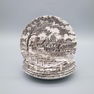 Puede incluir: Una pila de cuatro platos de cerámica blanca con un diseño marrón. Los platos presentan una escena detallada de un paisaje con un carruaje tirado por caballos, árboles y un edificio. Los platos están apilados sobre una superficie azul claro.