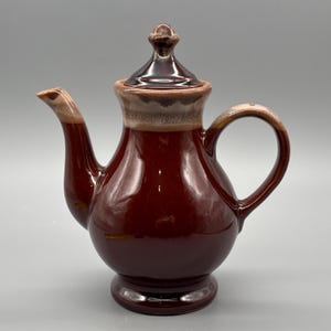 May include: A glossy, maroon ceramic teapot with a matching lid. The teapot features a curved spout, a rounded handle, and a base. The rim and handle have a tan and brown colour. The teapot is designed for serving tea.