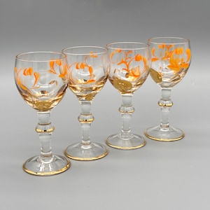 Puede incluir: Cuatro copas de vino transparentes con bordes y tallos dorados. Cada copa presenta un diseño floral naranja y un medallón dorado cerca de la base. Las copas están dispuestas sobre una superficie gris claro.