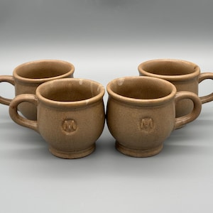 May include: Four beige ceramic mugs with rounded bodies and small handles. Each mug has a small, embossed design near the base. The mugs are arranged in a group, showcasing their simple, rustic design. These mugs are suitable for coffee or tea.