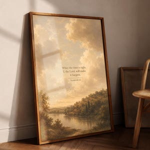 May include: A rectangular framed wall art print featuring a landscape painting of a river, rolling hills, and a cloudy sky. The text reads: When the time is right, I, the Lord, will make it happen. Isaiah 60:22. The frame is made of medium-toned wood.