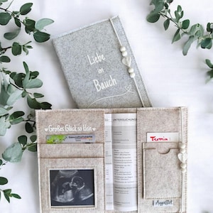 Könnte beinhalten: Ein hellgraues Filz-Schwangerschaftstagebuch und ein beiger Filz-Organizer. Das Tagebuch trägt den Text "Liebe im Bauch" und ist mit weißen Perlen und Herzen verziert. Der Organizer hat Taschen für Dokumente, ein Ultraschallfoto und eine Karte.