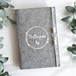 Op de afbeelding: Grijze vilten hoes met een wit cirkelvormig ontwerp met de tekst "Mutterpass" met twee voetafdrukken in de cirkel. Een wit hart en kralen zijn aan de hoes bevestigd.