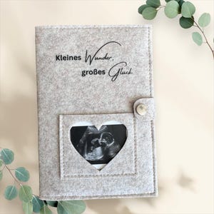 Op de afbeelding: Lichtgrijs vilten baby echo fotoalbum met een hartvormig venster. Het album heeft de Duitse tekst "Kleines Wunder, großes Glück" in zwart schrift. Een zilveren drukknoop sluit het album af.
