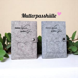 Könnte beinhalten: Zwei graue Filz-Mutterpasshüllen mit herzförmigem Ausschnitt und Druckknopfverschluss. Auf der Vorderseite steht der Text "Kleines Wunder, großes Glück". Ein rosa Herz befindet sich neben dem Wort "Mutterpasshülle".