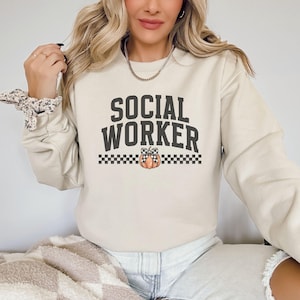 May include: A cream-colored sweatshirt with the text "SOCIAL WORKER" in black letters with a checkered border and a pumpkin with a bow in the center.