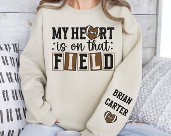 Könnte beinhalten: Ein cremefarbener Pullover mit dem Text "My heart is on that FIELD" in braunen Buchstaben. Der Text "BRIAN CARTER" befindet sich auf dem linken Ärmel mit einer braunen Fußballherzgrafik.