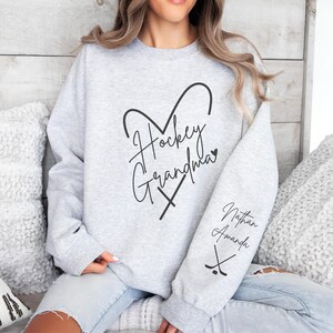 K&ouml;nnte beinhalten: Grauer Sweatshirt mit dem Text "Hockey Grandma" in Herzform mit Hockeyschl&auml;gern. Der &Auml;rmel hat den Text "Nathan Amanda" mit Hockeyschl&auml;gern.