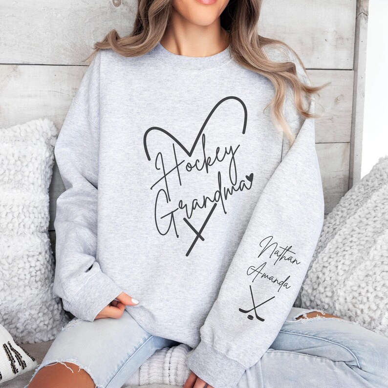 K&ouml;nnte beinhalten: Grauer Sweatshirt mit dem Text "Hockey Grandma" in Herzform mit Hockeyschl&auml;gern. Der &Auml;rmel hat den Text "Nathan Amanda" mit Hockeyschl&auml;gern.