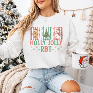 May include: White sweatshirt with a festive Christmas design. The design includes a gingerbread man, a Christmas tree, and a snowman, with the words "HOLLY JOLLY RBT" in colorful letters. The sweatshirt is paired with blue jeans.