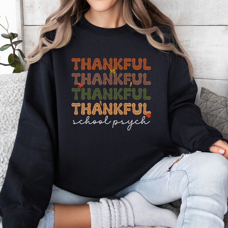 Puede incluir: Sudadera negra con el texto "THANKFUL" repetido cuatro veces en diferentes colores y "school psych" escrito debajo. El texto est&aacute; rodeado de peque&ntilde;os dise&ntilde;os de hojas.