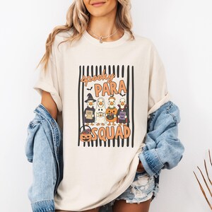 May include: A cream-colored t-shirt featuring a graphic of four ducks dressed in Halloween costumes, including a witch, ghost, pumpkin, and vampire. The text reads SPOOKY PARA SQUAD, set against a background of vertical black stripes and a jack-o'-lantern.