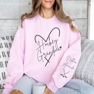 K&ouml;nnte beinhalten: Rosafarbener Pullover mit dem Text "Hockey Grandma" in Herzform mit Hockeyschl&auml;gern. Der &Auml;rmel hat den Text "Nathan Amanda" und eine Grafik mit einem Hockeyschl&auml;ger.