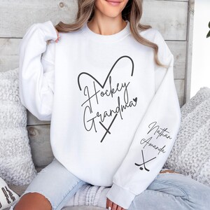 K&ouml;nnte beinhalten: Wei&szlig;er Pullover mit dem Text "Hockey Grandma" in Herzform mit gekreuzten Hockeyschl&auml;gern. Der &Auml;rmel hat den Text "Nathan Amanda" mit gekreuzten Hockeyschl&auml;gern.