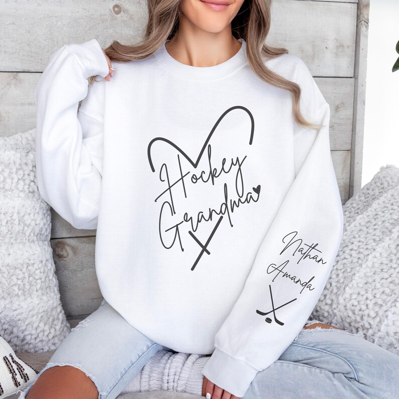 K&ouml;nnte beinhalten: Wei&szlig;er Pullover mit dem Text "Hockey Grandma" in Herzform mit gekreuzten Hockeyschl&auml;gern. Der &Auml;rmel hat den Text "Nathan Amanda" mit gekreuzten Hockeyschl&auml;gern.