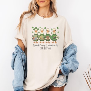 Op de afbeelding: Crèmekleurig T-shirt met een afbeelding van vier cartoon-eenden, elk met verschillende items. De eenden dragen groene outfits en zijn omringd door klavers. De tekst op het shirt luidt "Speech Goals & Shamrocks SLP EDITION."
