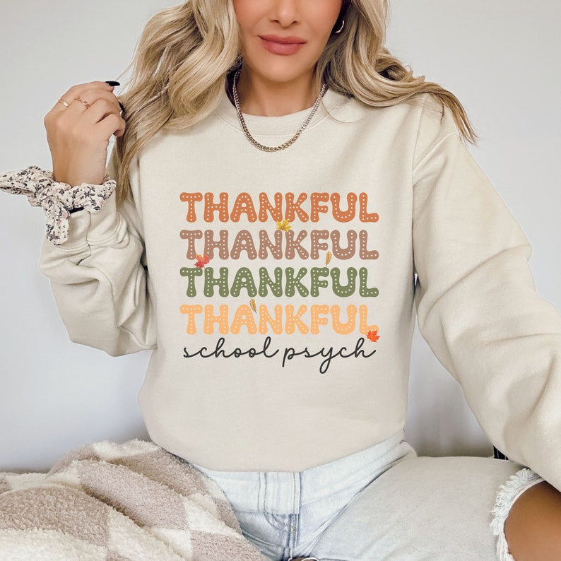 Puede incluir: Una sudadera beige con el texto "THANKFUL" repetido cuatro veces en diferentes colores y fuentes. El texto "school psych" est&aacute; escrito debajo del texto repetido. La sudadera tiene un tema oto&ntilde;al con peque&ntilde;os dise&ntilde;os de hojas.