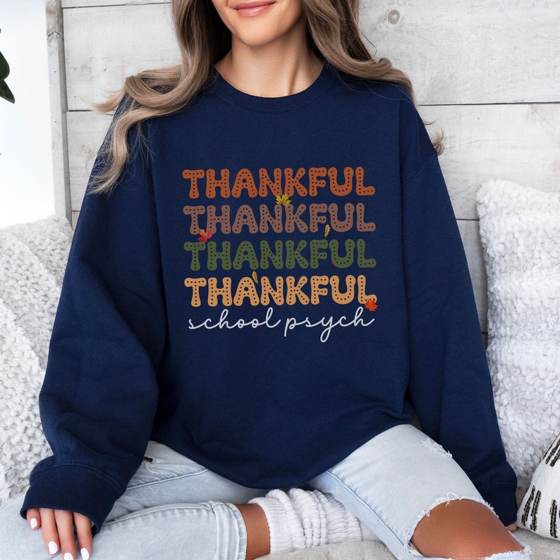 Puede incluir: Sudadera azul marino con el texto "THANKFUL" repetido cuatro veces en diferentes colores y "school psych" escrito debajo. El texto est&aacute; rodeado de peque&ntilde;os dise&ntilde;os de hojas.