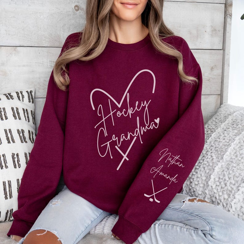 K&ouml;nnte beinhalten: Burgunderfarbener Pullover mit dem Text "Hockey Oma" in Herzform mit Hockeyschl&auml;gern. Der &Auml;rmel hat den Text "Nathan Amanda" mit Hockeyschl&auml;gern.