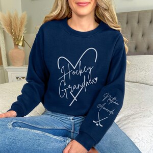 K&ouml;nnte beinhalten: Navyblauer Sweatshirt mit einem wei&szlig;en Herzdesign, auf dem "Hockey Grandma" und zwei gekreuzte Hockeyschl&auml;ger stehen. Der Sweatshirt hat einen wei&szlig;en Text auf dem &Auml;rmel, der "Nathan Amanda" und eine gekreuzte Hockeyschl&auml;gergrafik zeigt.