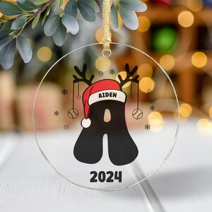 May include: A clear glass Christmas ornament with a black letter A wearing a red Santa hat with the name Aiden and the year 2024. The ornament is decorated with snowflakes and baseballs.