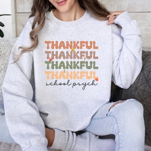 Puede incluir: Una sudadera gris con el texto "THANKFUL" repetido cuatro veces en diferentes colores. El texto "school psych" est&aacute; escrito debajo del texto repetido.