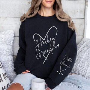 K&ouml;nnte beinhalten: Schwarzer Pullover mit einem wei&szlig;en Grafikdesign, auf dem "Hockey Grandma" mit einem Herzen und Hockeyschl&auml;gern steht. Der &Auml;rmel hat den Text "Nathan Amanda" und eine Grafik mit einem Hockeyschl&auml;ger.