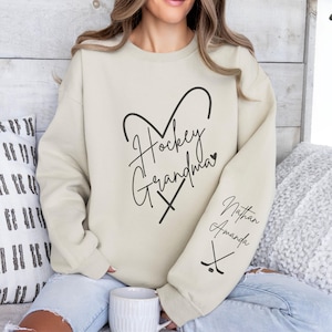 K&ouml;nnte beinhalten: Ein hellbeiger Pullover mit dem Text "Hockey Grandma" in Herzform mit Hockeyschl&auml;gern. Der &Auml;rmel hat den Text "Nathan Amanda" mit Hockeyschl&auml;gern.