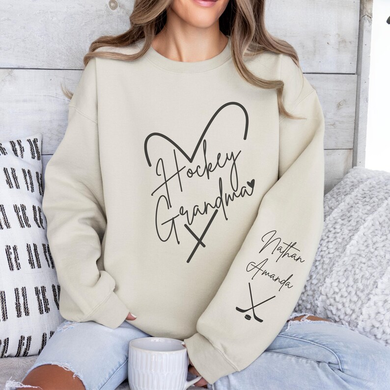 K&ouml;nnte beinhalten: Ein hellbeiger Pullover mit dem Text "Hockey Grandma" in Herzform mit Hockeyschl&auml;gern. Der &Auml;rmel hat den Text "Nathan Amanda" mit Hockeyschl&auml;gern.
